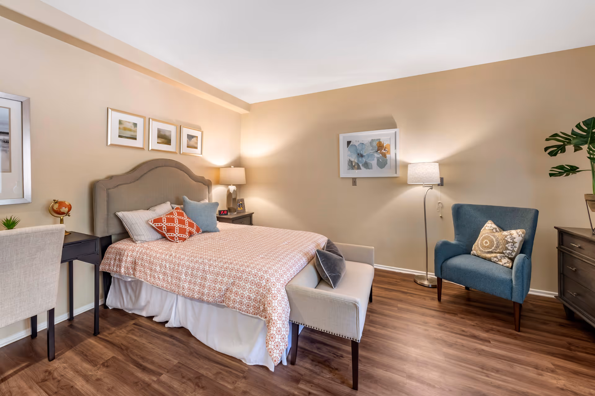 A cozy bedroom with a bed featuring a patterned bedspread and multiple pillows, a small bench at the foot of the bed, a blue armchair with a decorative pillow, a floor lamp, a dresser with a plant, and framed artwork on the walls. The room has wooden flooring and soft lighting.