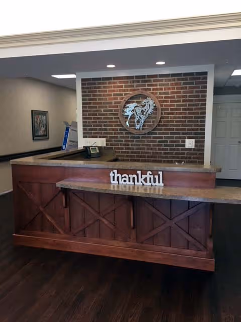 Reception desk area with a wooden counter and a decorative sign that says 'thankful'. Behind the desk is a brick wall with a circular wooden art piece featuring a horse design. The floor is dark wood, and there is a doorway and framed artwork visible in the background.