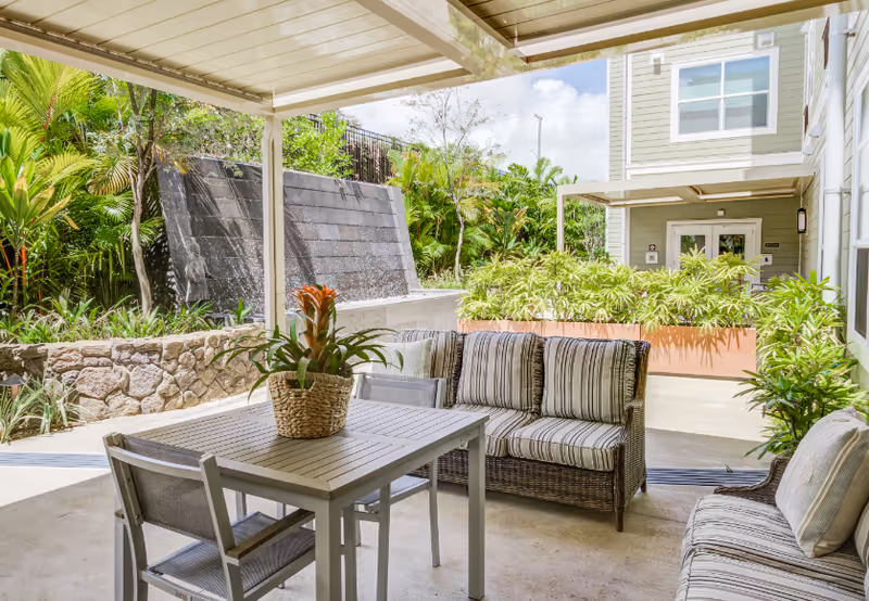 Covered outdoor patio area with a table and chairs, cushioned wicker seating, a potted plant on the table, and a stone water feature wall surrounded by lush greenery.