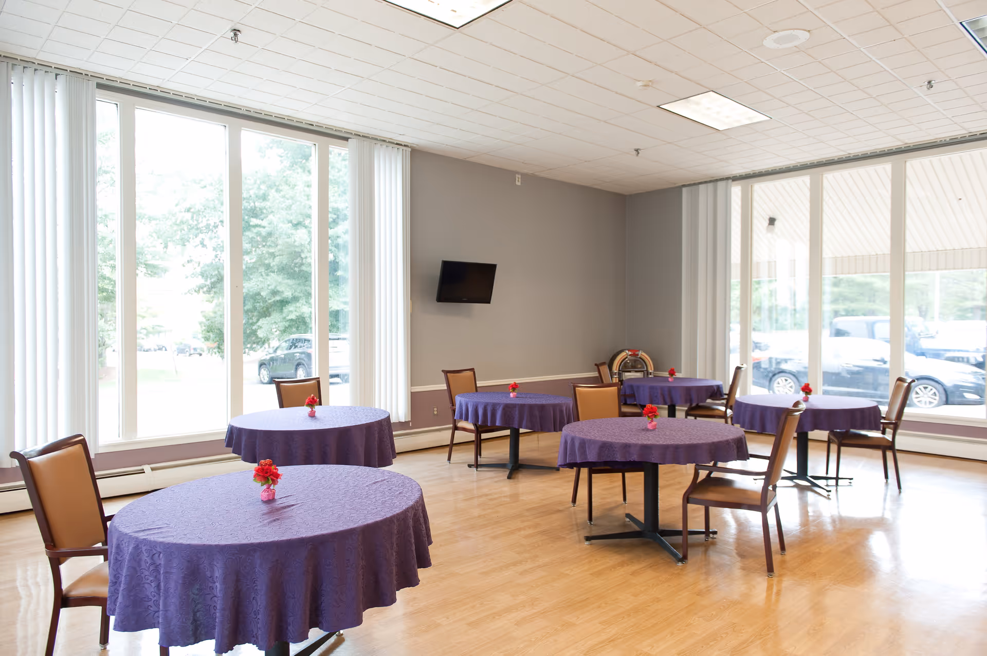 A bright dining room with several round tables covered in purple tablecloths, each adorned with a small red flower centerpiece. The room has large windows letting in natural light, wooden floors, and a wall-mounted TV. Chairs are arranged around the tables, and a jukebox is visible in the corner.