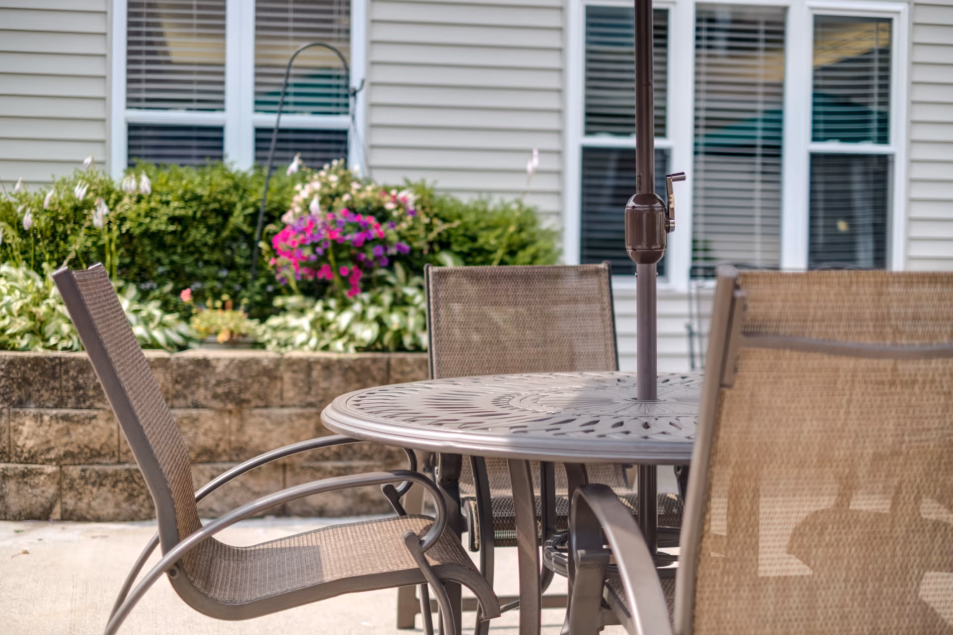 Outdoor patio area with a round metal table and four mesh chairs. Behind the patio is a raised garden bed with green shrubs and blooming flowers, and a building with beige siding and windows with white blinds.