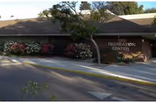 Exterior view of a single-story building labeled '1200 Recreation Center' with a sloped roof, surrounded by bushes and trees, and a paved driveway in front.