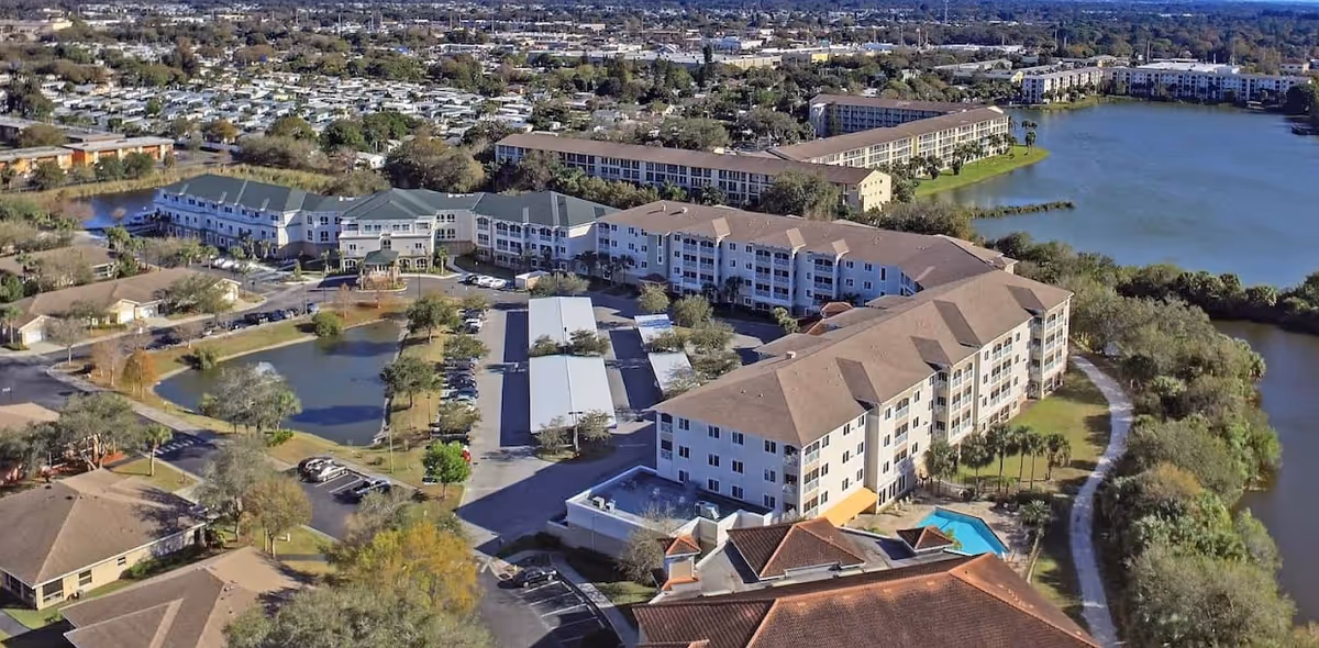 Aerial view of Water's Edge of Bradenton senior living facility showing multiple large residential buildings surrounding parking areas, ponds, and a swimming pool, with a lake and greenery in the background.