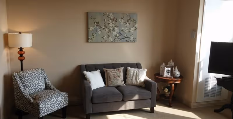 A cozy living room with a gray loveseat adorned with three decorative pillows, a patterned armchair, a wooden side table with decorative items including a photo frame and vases, a floor lamp with a white shade, and a flat-screen TV on a stand. A floral painting hangs on the beige wall above the loveseat.