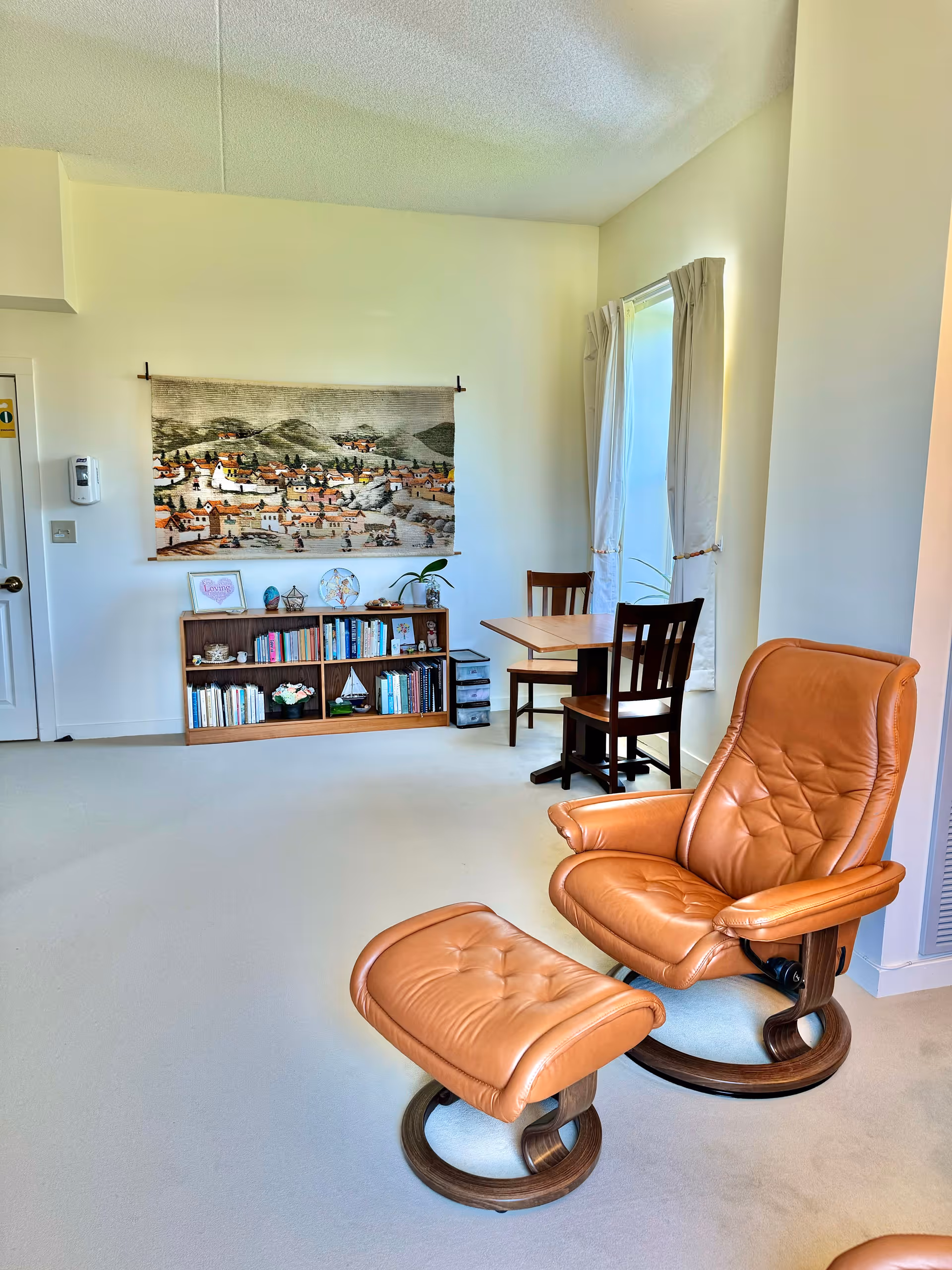 A cozy living area with a tan leather recliner and matching ottoman on a light carpeted floor. In the background, there is a wooden bookshelf filled with books and decorative items, a wall hanging depicting a village scene, and a small wooden table with two chairs near a window with white curtains.