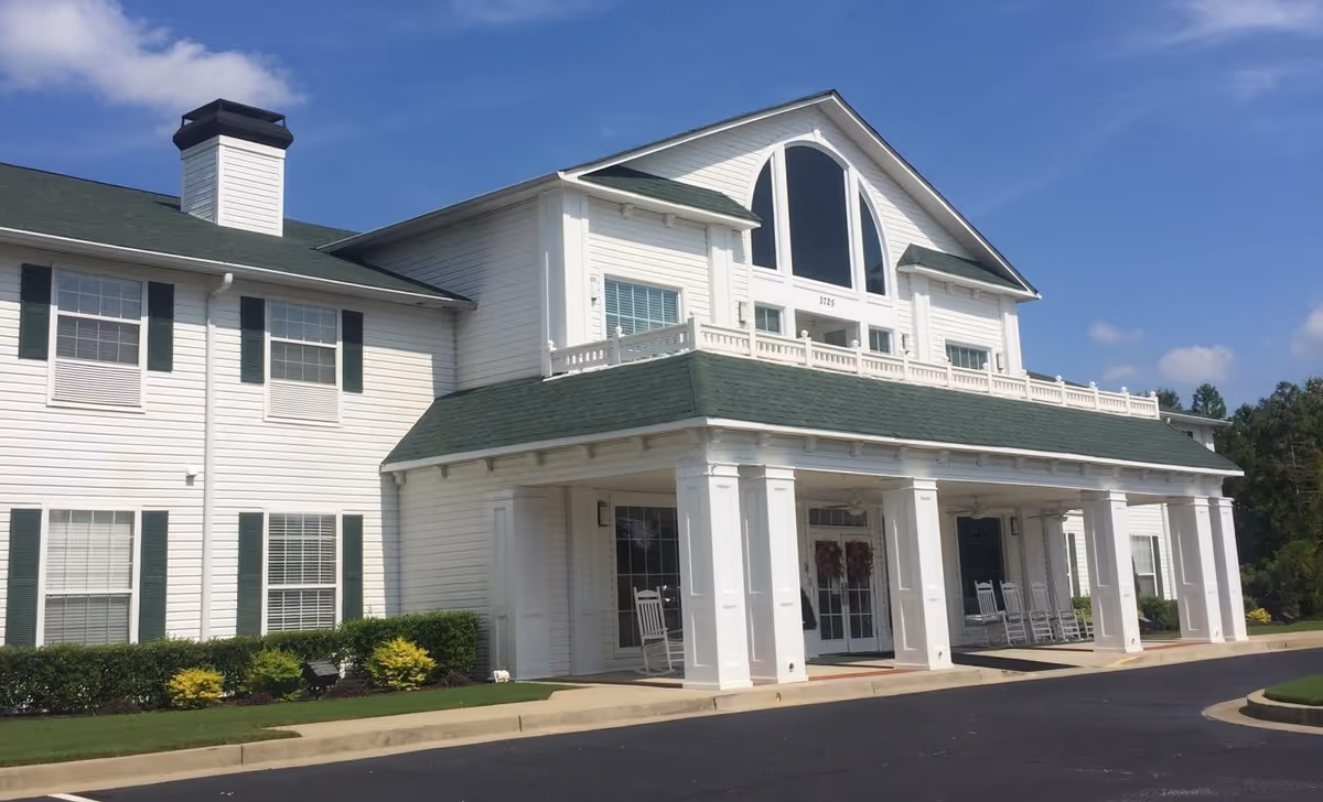 Front exterior view of a two-story senior living facility building with white siding, green shutters, and a green roof. The entrance features a covered porch with white columns and rocking chairs. The sky is clear and blue.