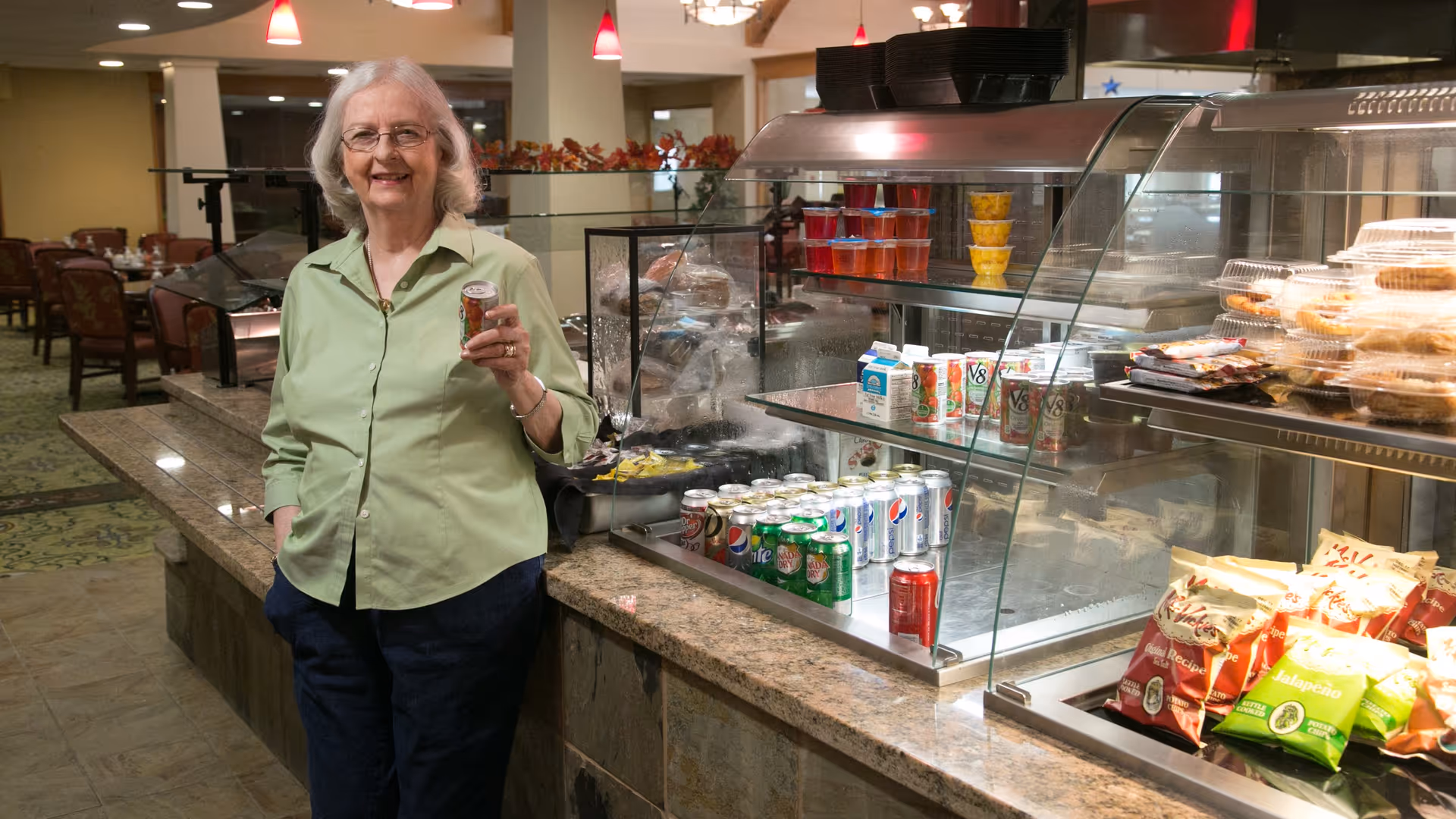 An elderly woman with gray hair and glasses wearing a light green shirt and dark pants stands smiling next to a food and beverage display counter in a dining area. The counter holds various canned drinks, juice boxes, snack bags, and packaged baked goods. The background shows tables and chairs in a well-lit dining room.