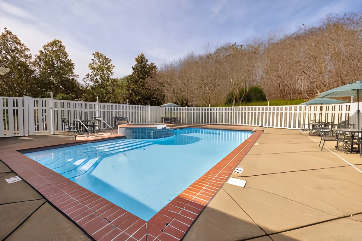 Outdoor swimming pool with brick coping, surrounding patio seating and a white picket fence under a clear sky.