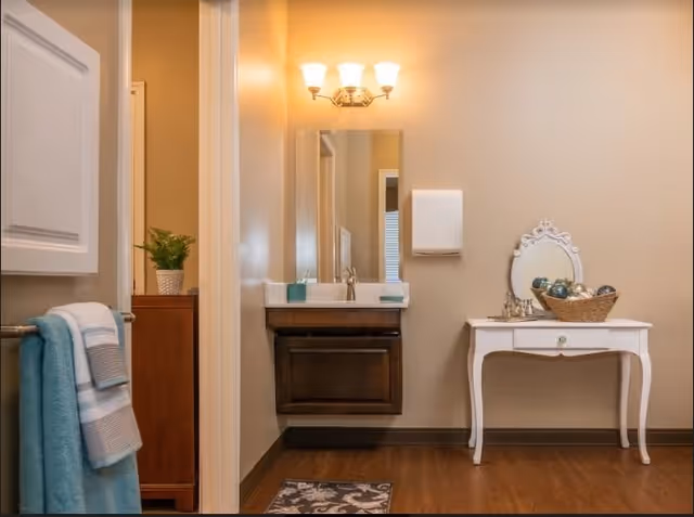 Interior view of a bathroom area with a towel rack holding blue and white towels on the left, a wooden cabinet with a potted plant, a wall-mounted sink with a mirror and light fixture above it, a paper towel dispenser on the wall, and a white vanity table with a decorative mirror and a basket of decorative balls on the right.
