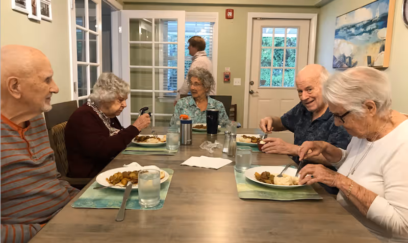 Five elderly people sitting around a dining table eating a meal together in a well-lit room with a door and windows in the background. One person is walking past the door in the background.