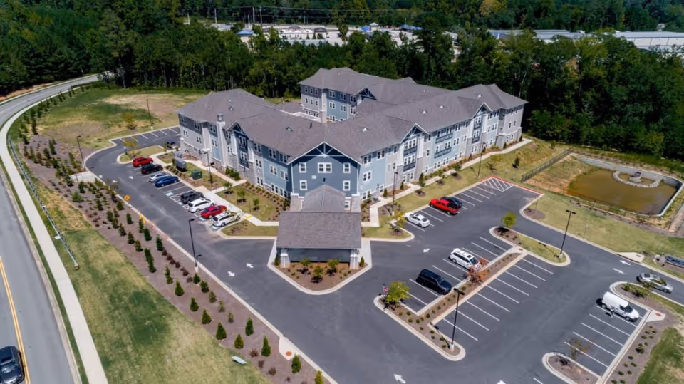 Aerial view of a large, three-story senior living facility building with a gray roof and blue-gray exterior walls, surrounded by a parking lot with several cars parked. The building is situated near a road and bordered by trees and greenery, with a small pond visible on the right side.
