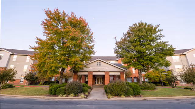 Front exterior view of Harding Place Retirement facility with a central entrance flanked by two large trees showing autumn foliage, surrounded by well-maintained bushes and grass.