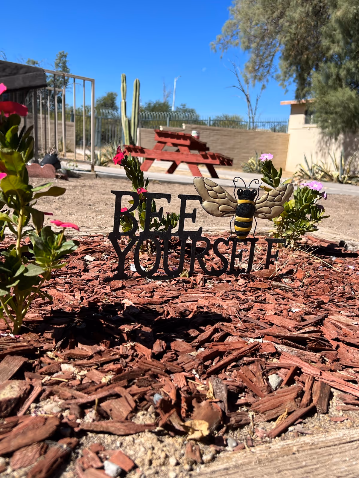 Decorative garden bed featuring a 'Bee Yourself' sign with a bee ornament and a red picnic table in the background under a clear blue sky.