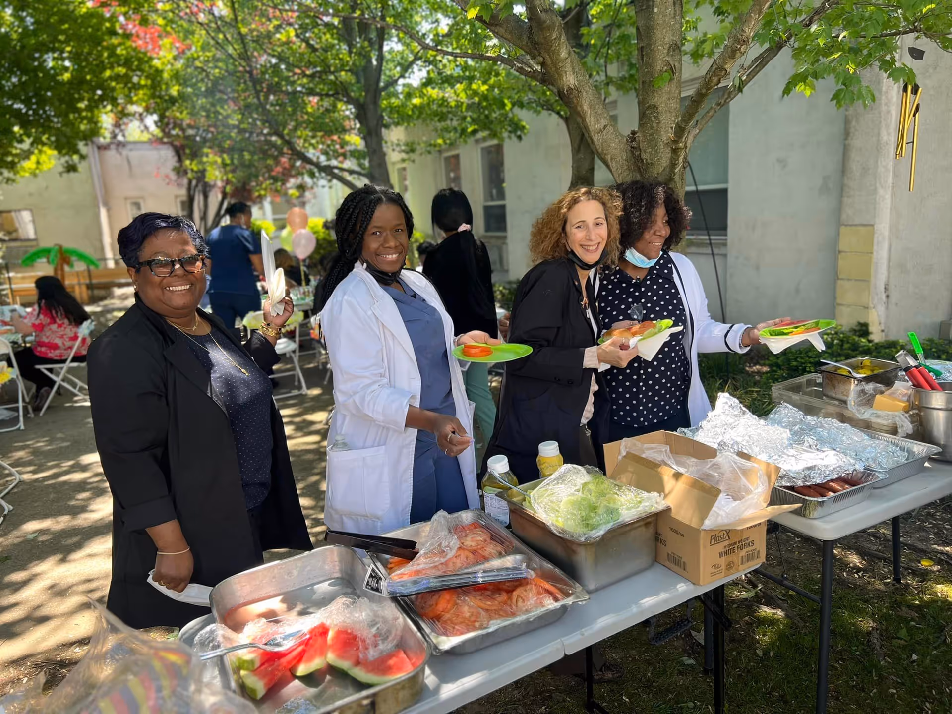 Four women standing outdoors behind a table with food trays and plates, smiling and holding plates with food. The setting appears to be a garden or patio area with trees and other people seated at tables in the background.