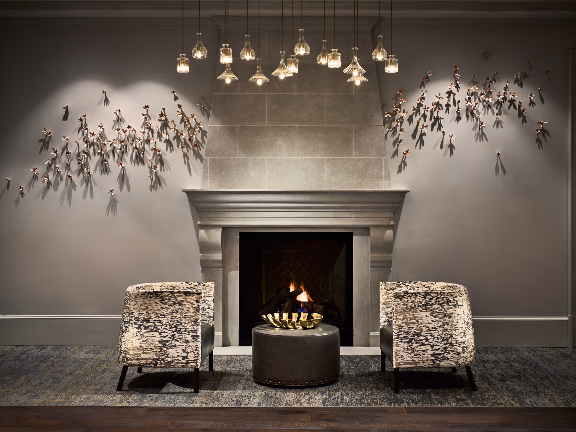 A cozy seating area with two patterned armchairs facing a lit fireplace with a stone mantel. Above the fireplace hangs a modern chandelier with multiple glass pendant lights. Decorative wall art featuring small metallic figures is displayed on both sides of the fireplace on a gray wall. A round ottoman with a decorative bowl sits between the chairs on a textured rug.