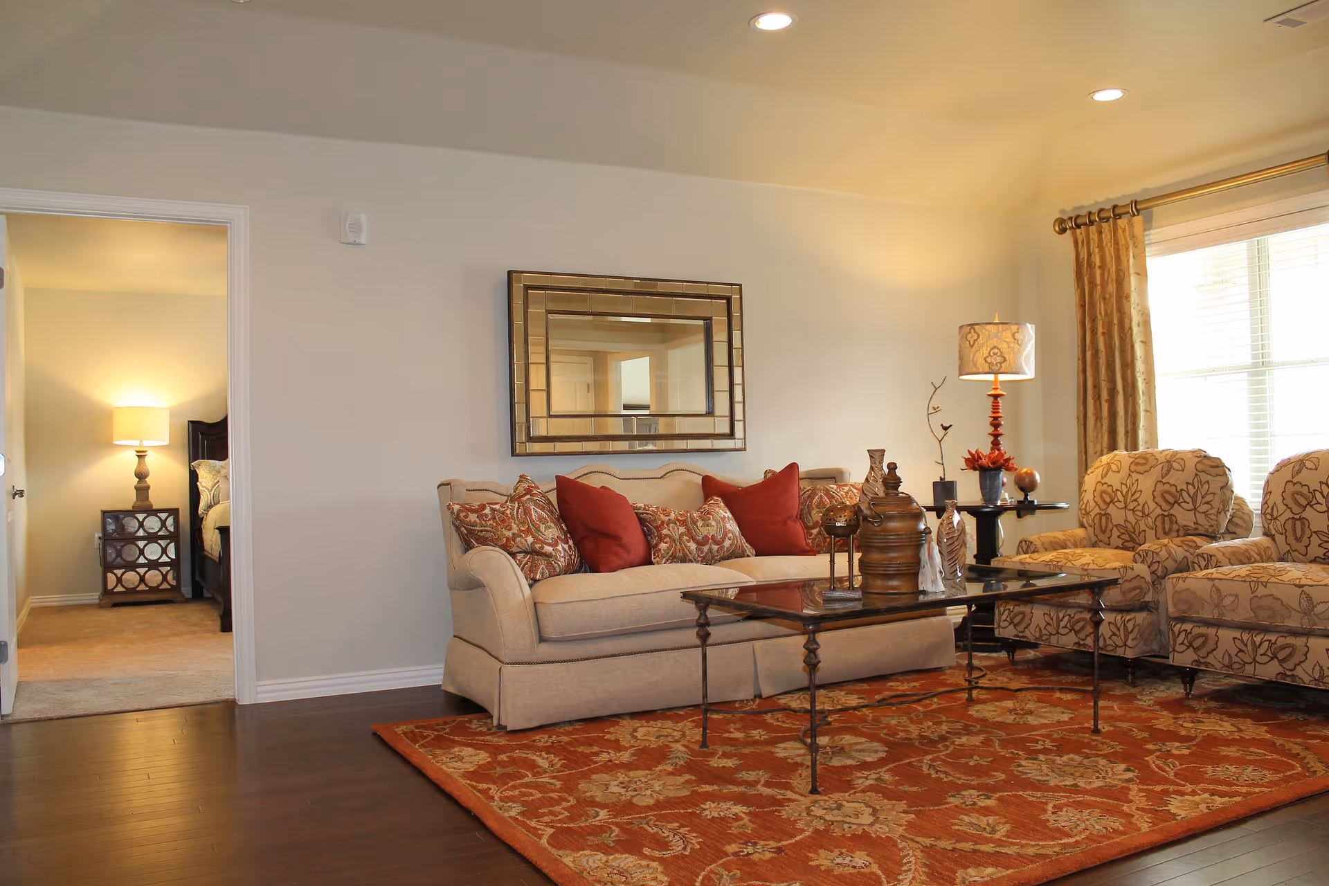 A cozy living room with a beige sofa adorned with patterned and solid red cushions, two matching floral armchairs, a glass coffee table with decorative items, a large ornate mirror on the wall, and a window with beige curtains letting in natural light. In the background, an open doorway reveals a bedroom with a lit table lamp on a nightstand.