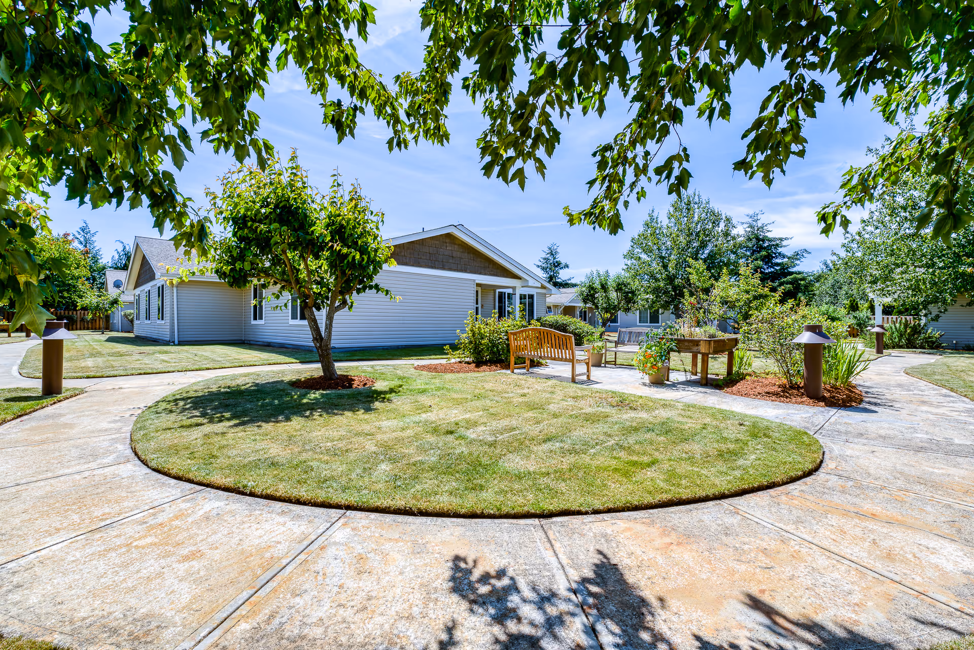 A sunny outdoor courtyard area at San Juan Villa Memory Care featuring a circular concrete walkway surrounding a grassy area with a small tree in the center. There are benches and a raised planter with flowers, along with various trees and shrubs around the perimeter. The sky is clear and blue.