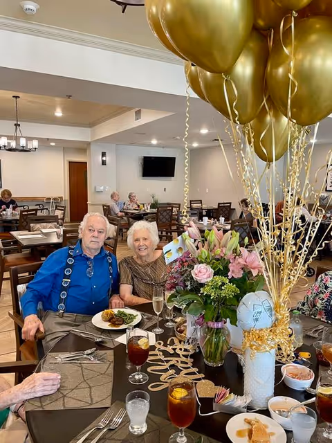 An elderly couple sitting at a dining table in a senior living facility dining room. The table is decorated with a large bouquet of flowers, gold balloons, and various drinks and plates of food. Other residents are seated at tables in the background.