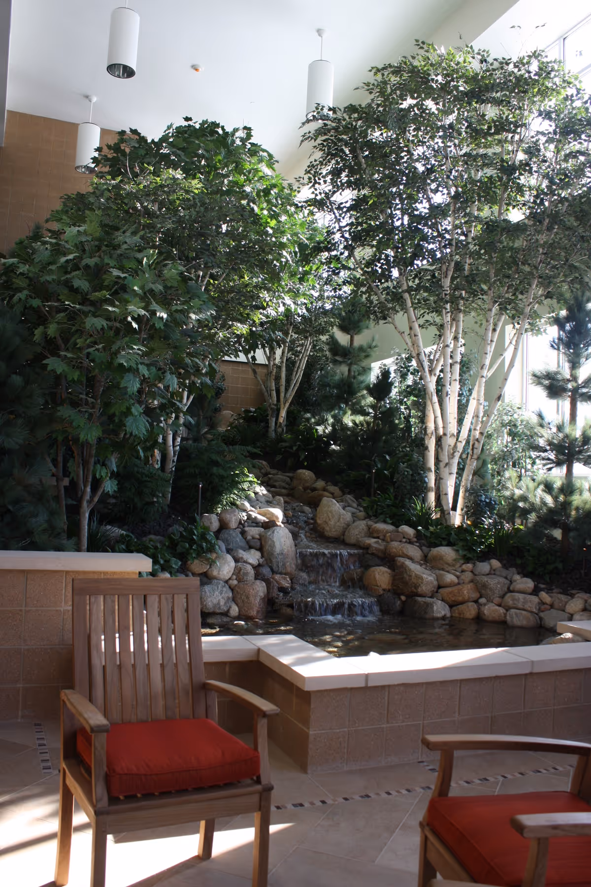 Indoor garden area with a small rock waterfall surrounded by lush green trees and plants. Two wooden chairs with red cushions are positioned near the water feature. The space is well-lit with natural light coming through large windows.
