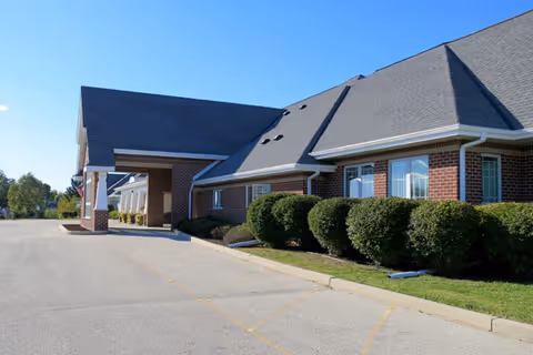 Exterior view of a single-story brick building with a gray shingled roof, featuring a covered entrance and neatly trimmed bushes along the side. The building is situated next to a paved driveway under a clear blue sky.