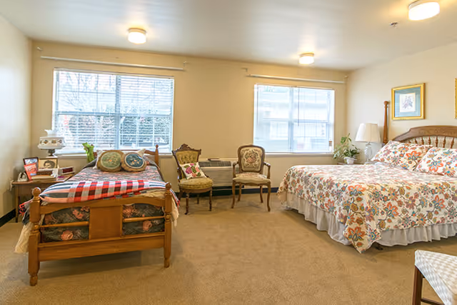 A bright bedroom with two beds, one single bed with a red and white checkered blanket and decorative pillows, and one larger bed with floral bedding. There are two windows letting in natural light, two vintage-style chairs between the beds, a nightstand with a lamp and picture frames, and a framed picture on the wall above the larger bed.