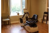 A room with wooden flooring featuring a recumbent exercise bike positioned near a window with beige curtains. There are two chairs against the walls, a small trash bin, and a potted plant on the windowsill.
