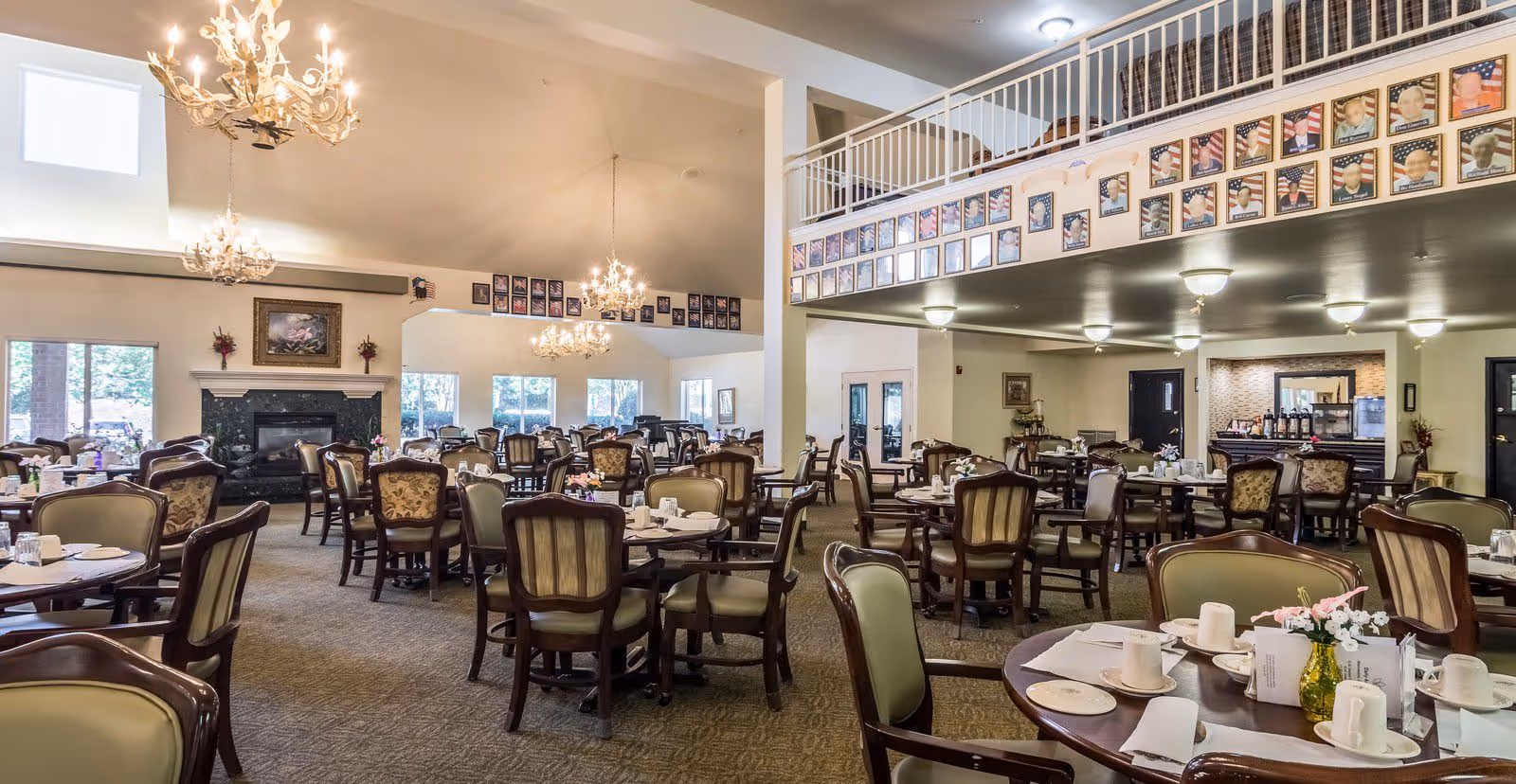 A spacious dining room with multiple round tables and cushioned chairs arranged neatly. Each table is set with white cups, plates, napkins, and small flower vases. The room features high ceilings with elegant chandeliers, large windows letting in natural light, and a fireplace with decorative flowers and a painting above it. There are framed photos displayed along the upper walls and a balcony area overlooking the dining space.