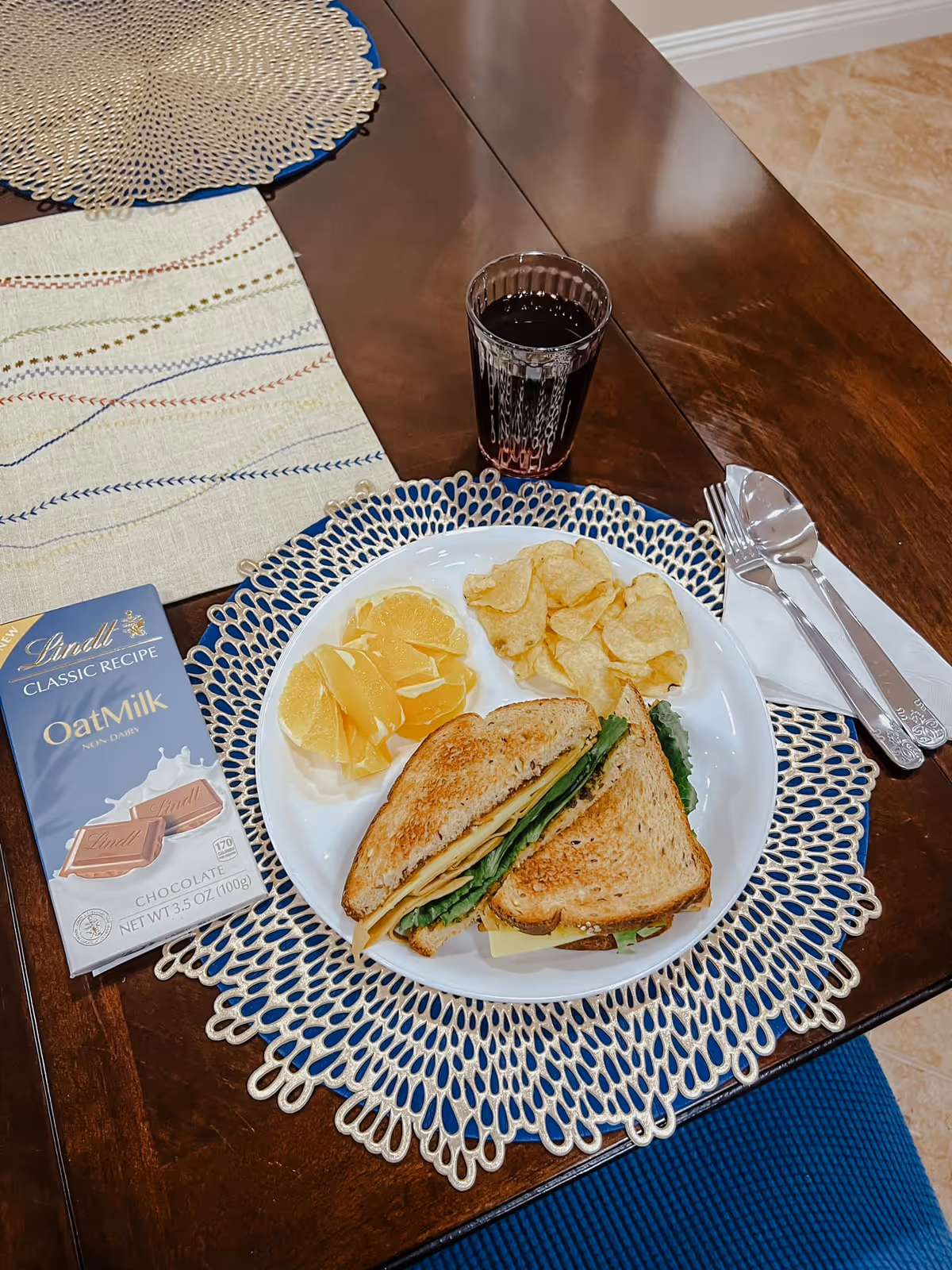 A wooden dining table set with a white plate containing a sandwich with lettuce and cheese, potato chips, and orange slices. Next to the plate is a glass of dark beverage and a bar of Lindt Oat Milk chocolate. The plate is placed on a decorative blue and beige placemat, and there is a set of silverware wrapped in a white napkin to the right.