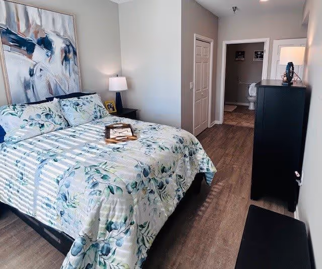 A bedroom with a bed covered in a floral patterned comforter and pillows. There is a nightstand with a lamp and a framed photo next to the bed. A large abstract painting hangs on the wall above the bed. The room has wood flooring and light-colored walls. In the background, there is an open doorway leading to a bathroom with a visible toilet and a dresser with a lamp on top.