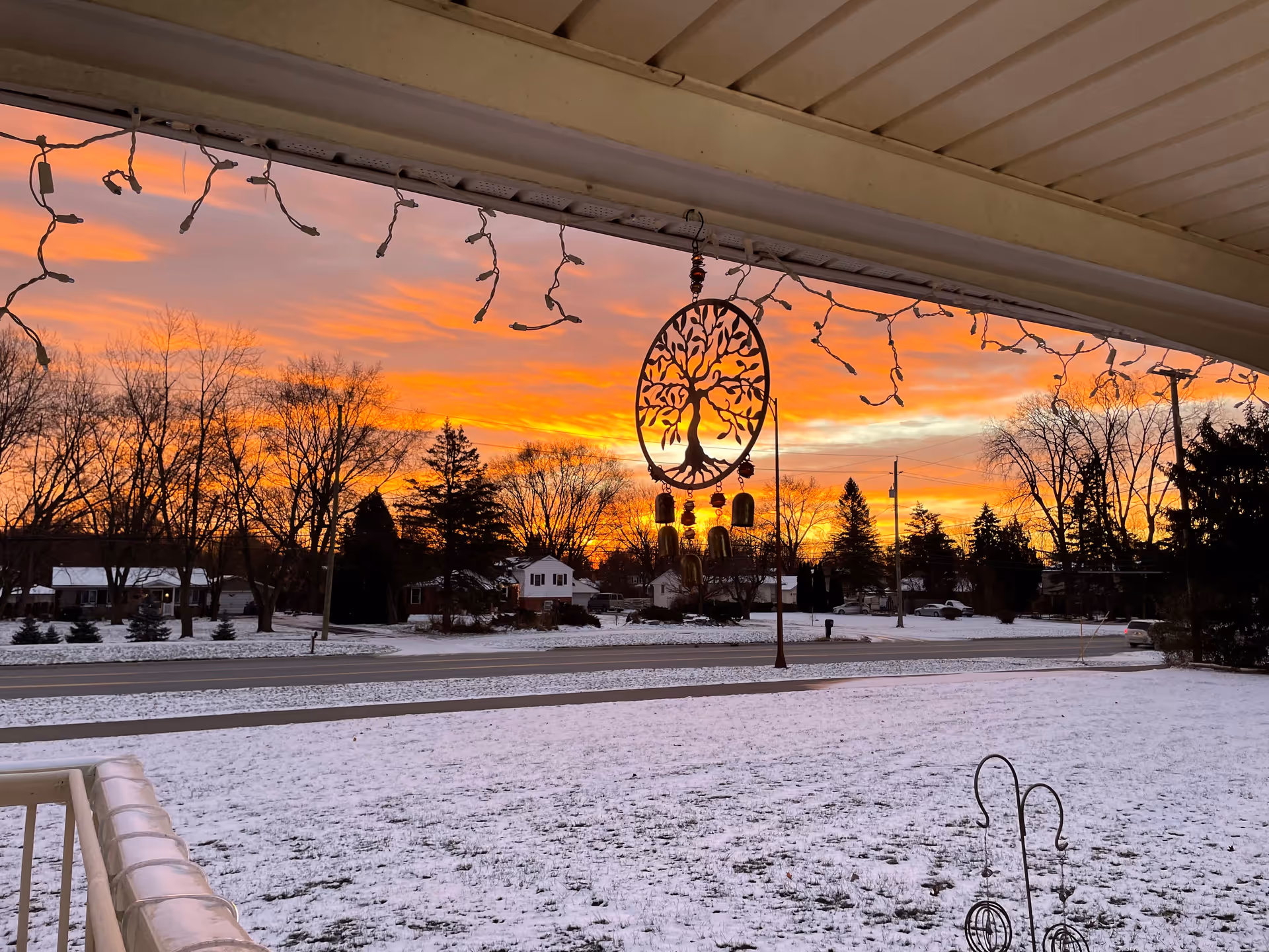 View from a covered porch overlooking a snowy yard and a street with houses and trees in the background during a vibrant orange and pink sunset. Hanging from the porch ceiling are string lights and a decorative wind chime with a tree design.