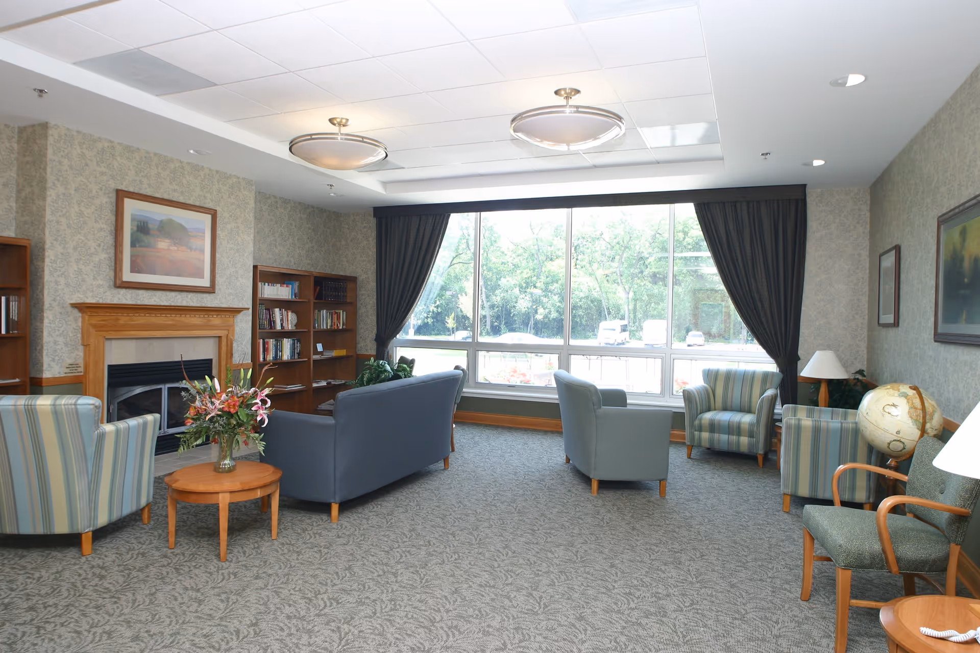 A spacious living room with patterned carpet, several upholstered chairs and sofas arranged around a wooden coffee table with a flower arrangement. There is a fireplace with a wooden mantle, bookshelves filled with books, large windows with dark curtains letting in natural light, and framed artwork on the walls.