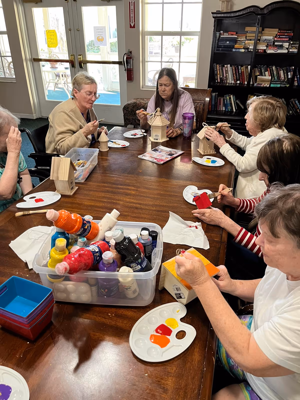 A group of elderly women sitting around a wooden table engaged in painting small wooden birdhouses. Various paint bottles and palettes with bright colors like red, yellow, and orange are on the table. In the background, there is a bookshelf filled with books and a glass door letting in natural light.