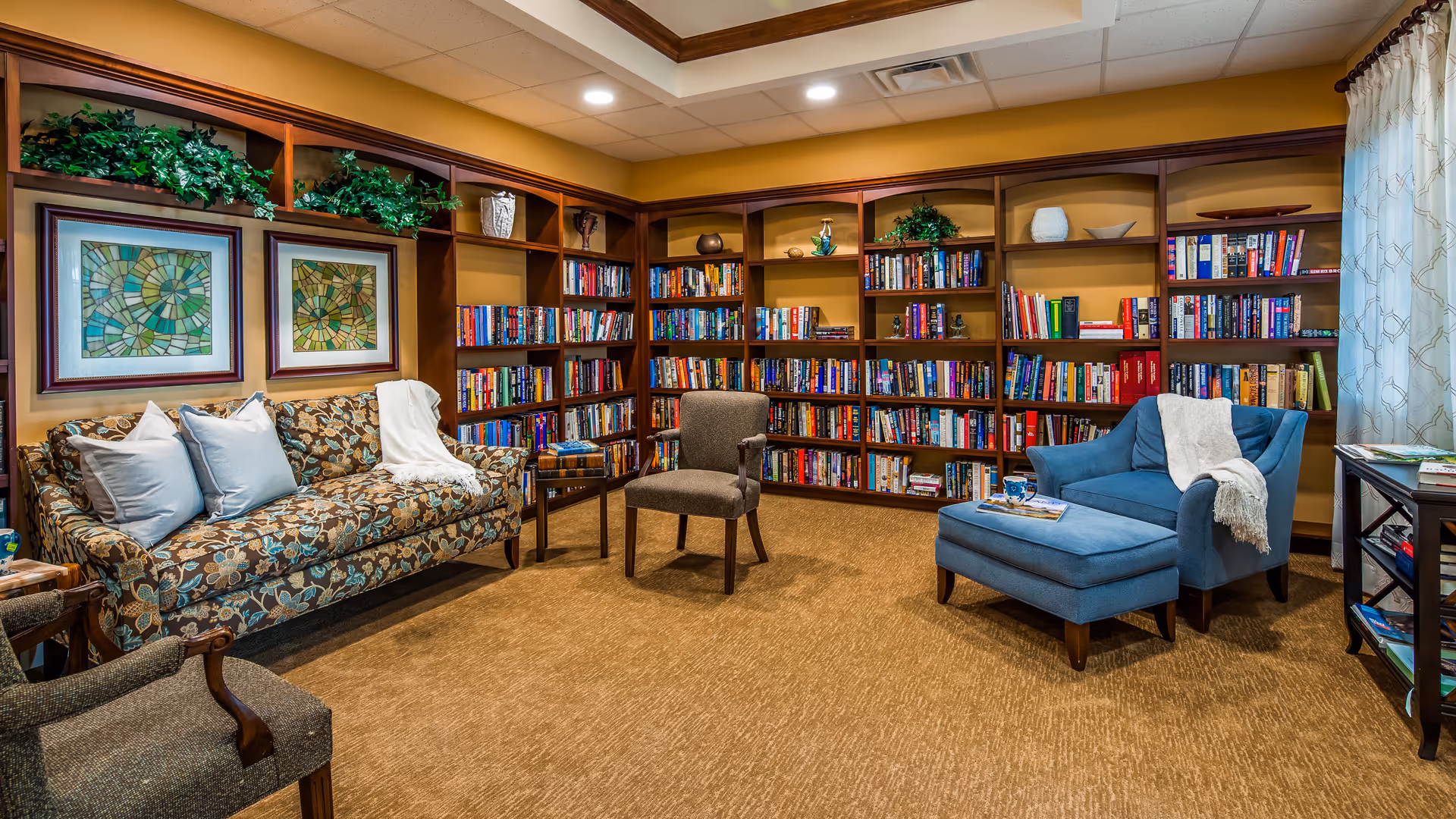 A cozy library room with wooden bookshelves filled with books along two walls. There is a floral patterned sofa with light blue pillows and a white throw blanket, a brown armchair, and a blue armchair with a matching ottoman and a white throw blanket. The room has warm lighting, framed artwork on the wall, and a window with light curtains.