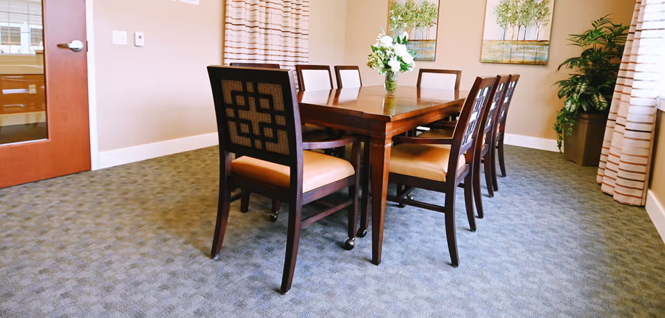 A dining room featuring a rectangular wooden table surrounded by chairs, a floral centerpiece, wall art, and potted plants.