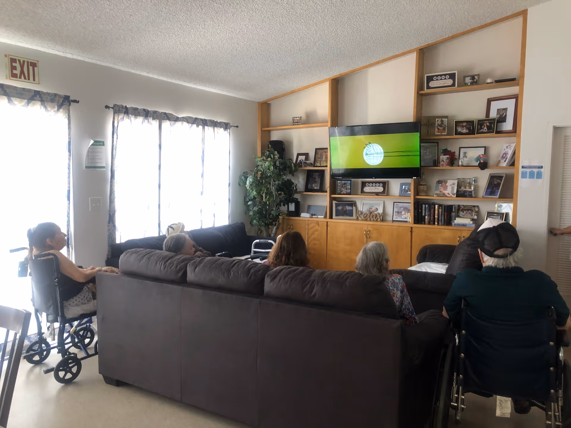 Several seniors, some in wheelchairs, sit on sofas facing a wall-mounted TV in a bright common living room with built-in shelving.