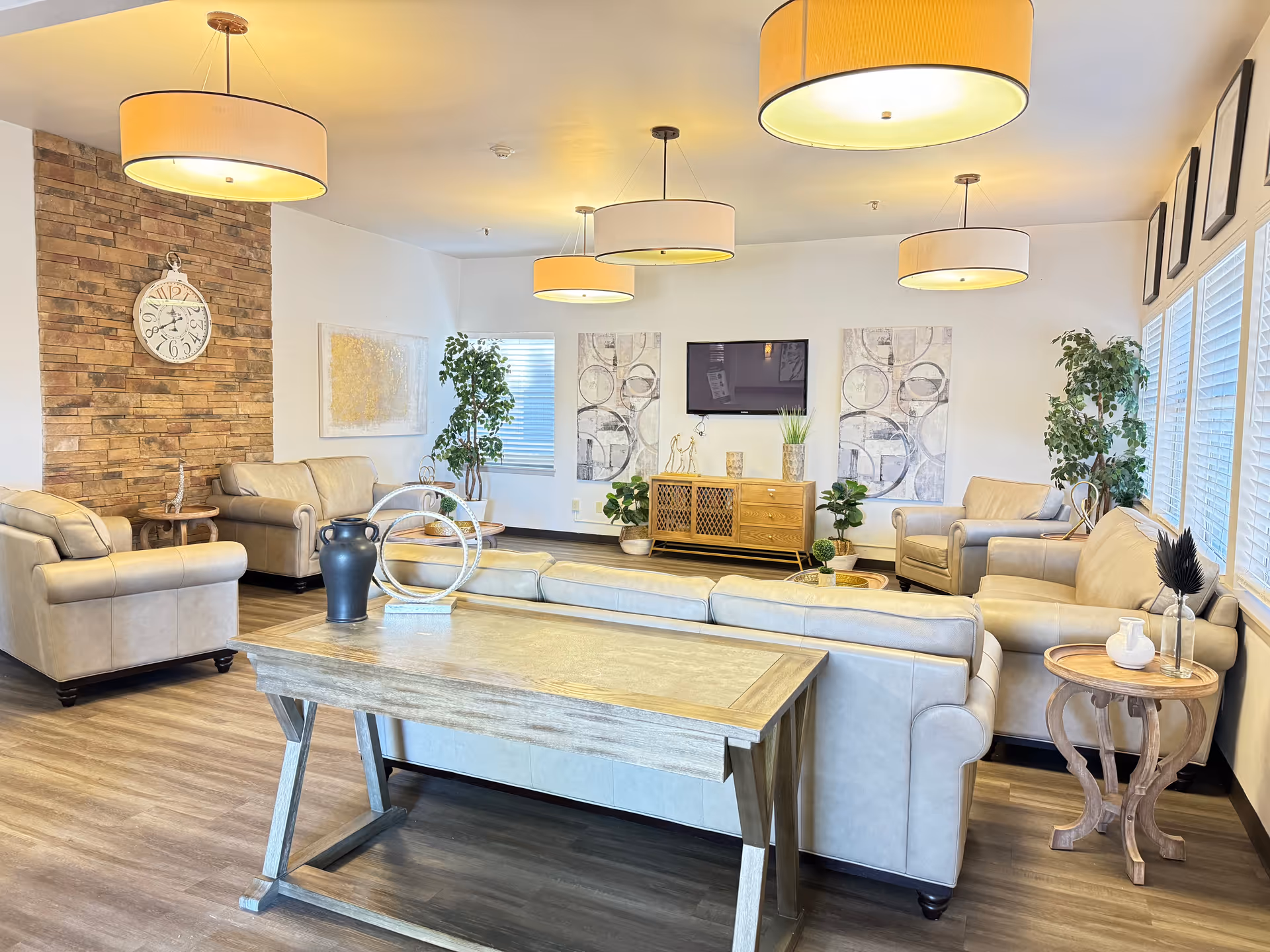 A bright and spacious living room area with beige leather sofas and armchairs arranged around a wooden coffee table and a wooden console with decorative items. The room features large windows with blinds, several potted plants, modern pendant lights hanging from the ceiling, a wall clock on a brick accent wall, and abstract artwork on the walls.