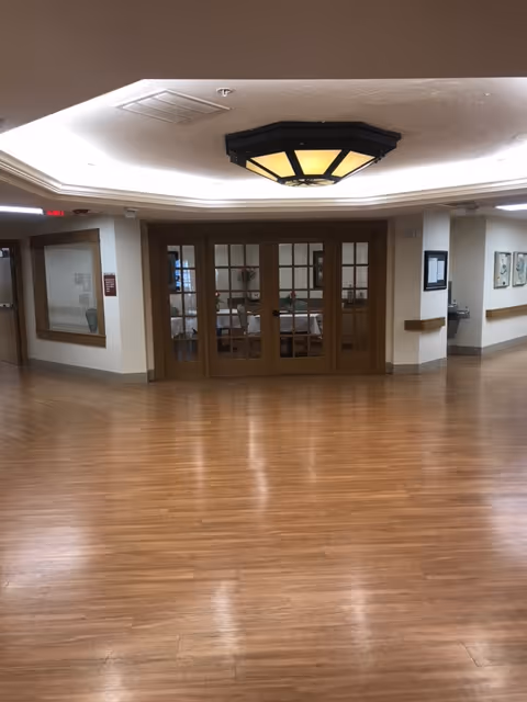 Interior hallway with wooden flooring leading to a room with large glass-paneled double doors. The ceiling has a decorative light fixture, and there are handrails along the walls. A water fountain and framed pictures are visible on the right side.