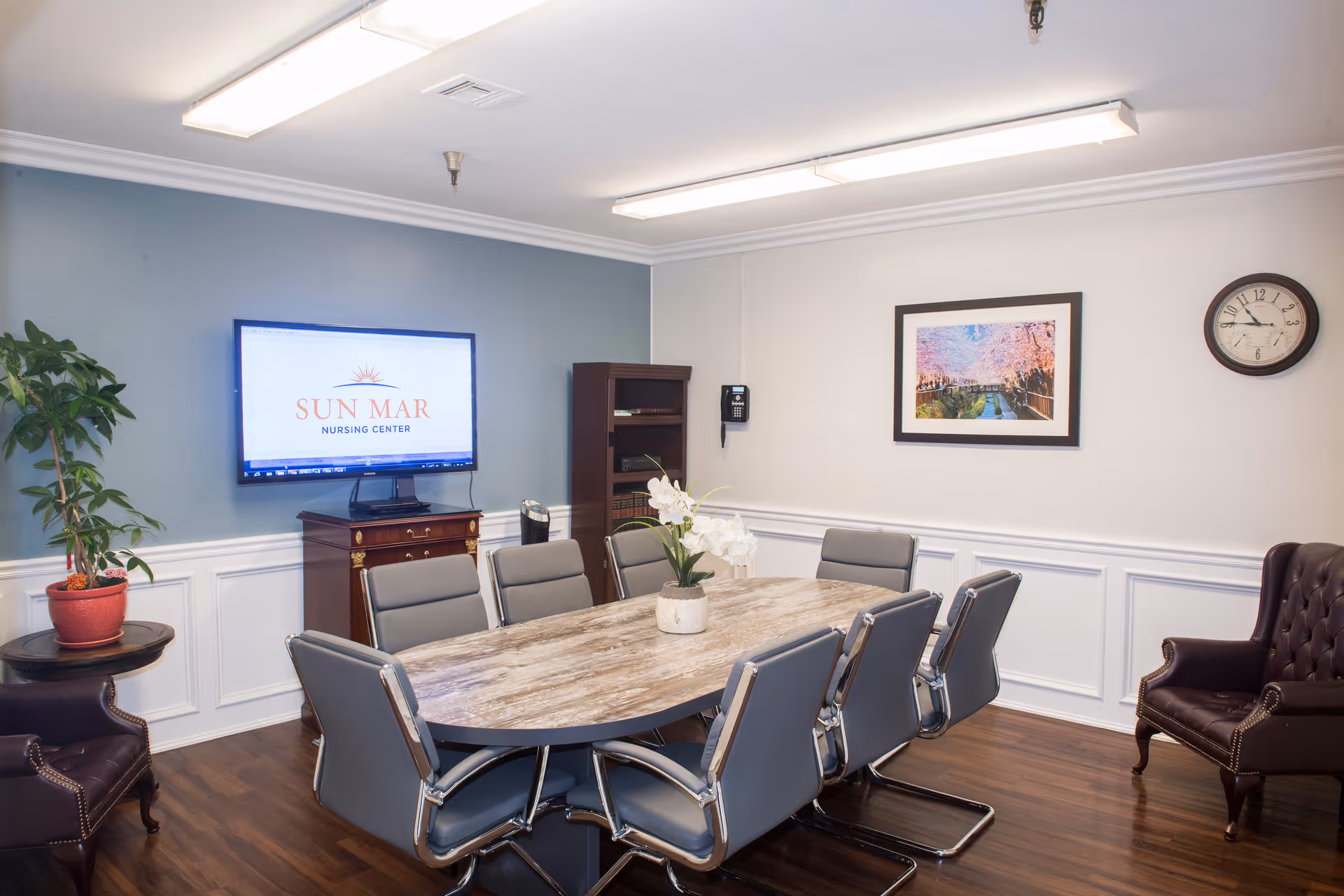 A conference room with a large oval wooden table surrounded by eight gray office chairs. On the left wall, a flat-screen TV displays the Sun Mar Nursing Center logo. There is a potted plant on a small round table and a dark wooden cabinet beneath the TV. The right wall features a framed picture of a scenic pathway with cherry blossoms and a round clock showing the time as 12:15. The room has white wainscoting, light blue and white walls, and wooden flooring.