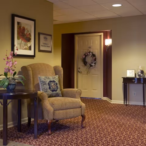 Upholstered armchair with a decorative pillow beside side tables in a carpeted hallway leading to a door adorned with a wreath.