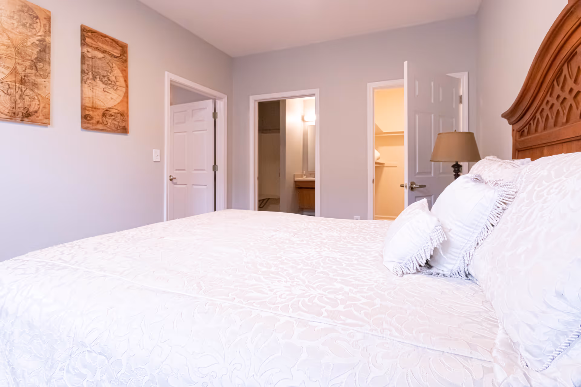 A bedroom with a large bed covered in white patterned bedding and pillows. The headboard is wooden with a carved design. On the wall to the left, there are two framed vintage map artworks. Three doorways are visible: one leading to a bathroom with a sink and mirror, another to a closet with shelves, and a third closed door. A lamp with a beige shade is on a nightstand next to the bed.