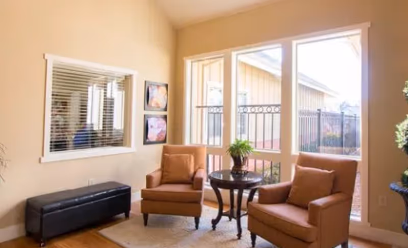 Sunlit sitting area with two upholstered armchairs and a small round table in front of large windows.