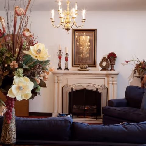 A cozy living room with a white fireplace adorned with decorative items including candles, a clock, and a vase with flowers. Above the fireplace hangs a framed picture reflecting a chandelier. In front of the fireplace are dark blue upholstered sofas, and to the left is a large floral arrangement in a decorative vase. A gold chandelier hangs from the ceiling.