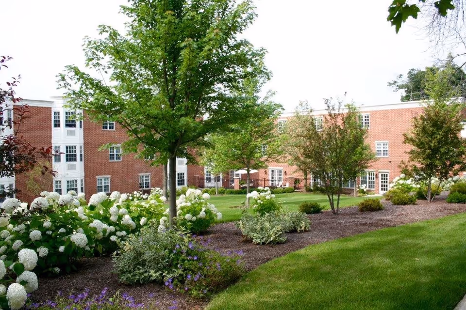 A landscaped outdoor garden area with green grass, flowering bushes, and several trees in front of a red brick senior living facility building with multiple windows and white trim.
