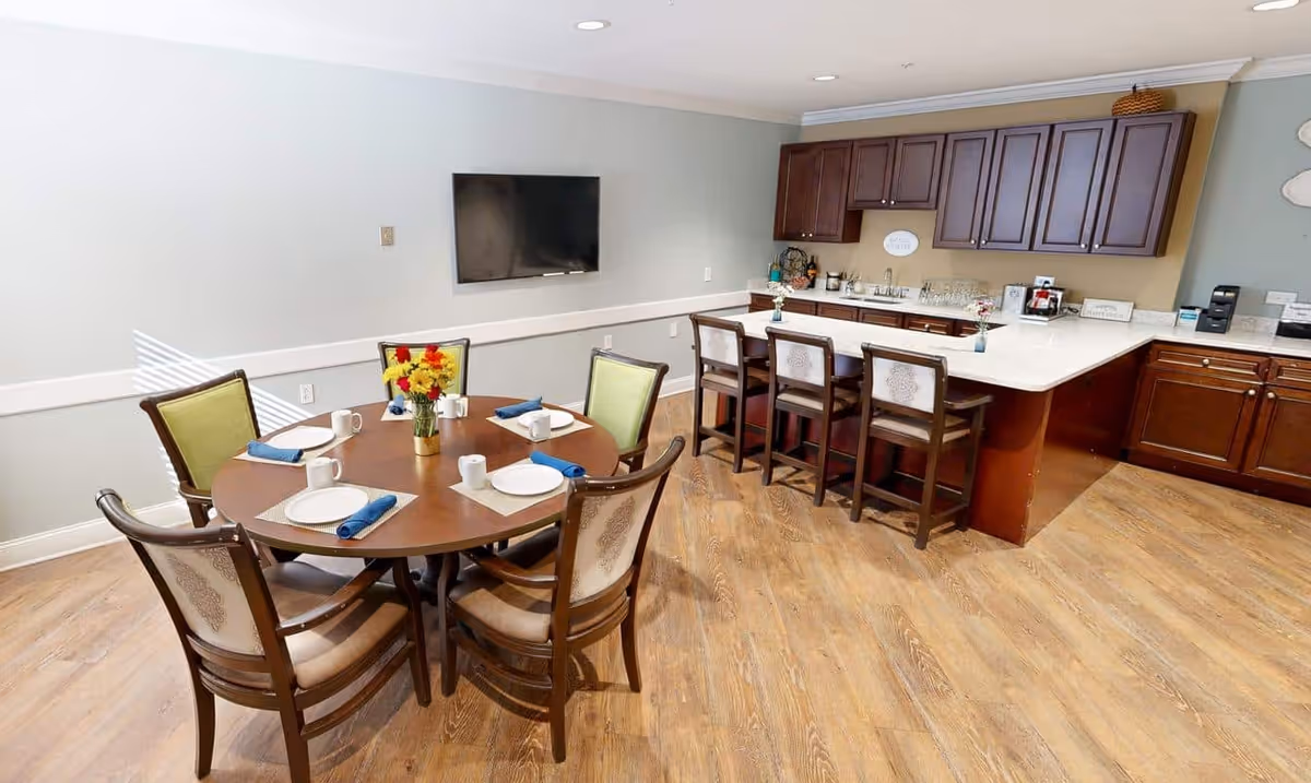 A dining area in Dunwoody Place featuring a round wooden table set with six chairs, each place set with a plate, mug, and blue napkin. A vase with colorful flowers is in the center of the table. In the background, there is a kitchen counter with four bar stools, dark wooden cabinets, and various kitchen items. A flat-screen TV is mounted on the light-colored wall.
