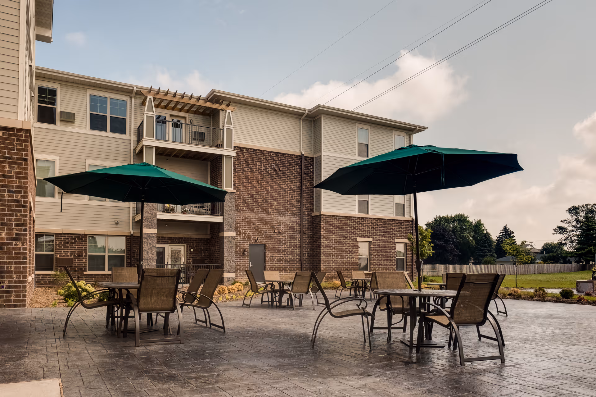 Outdoor patio area at Aspire Senior Living Kimberly with several tables and chairs under large green umbrellas, adjacent to a multi-story brick and siding building, with a grassy area and trees in the background under a partly cloudy sky.