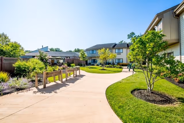 A sunny outdoor courtyard area at Arbor Terrace Middletown featuring a wide concrete pathway, green grass, small trees, and raised garden beds along a wooden fence. Residential buildings are visible in the background under a clear blue sky.