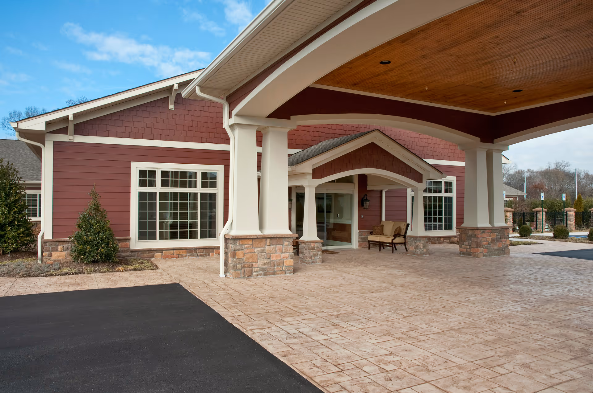Entrance area of a senior living facility with a covered drop-off zone supported by white columns with stone bases. The building exterior is red with white trim and large windows. There is a bench near the entrance door, and the sky is partly cloudy.