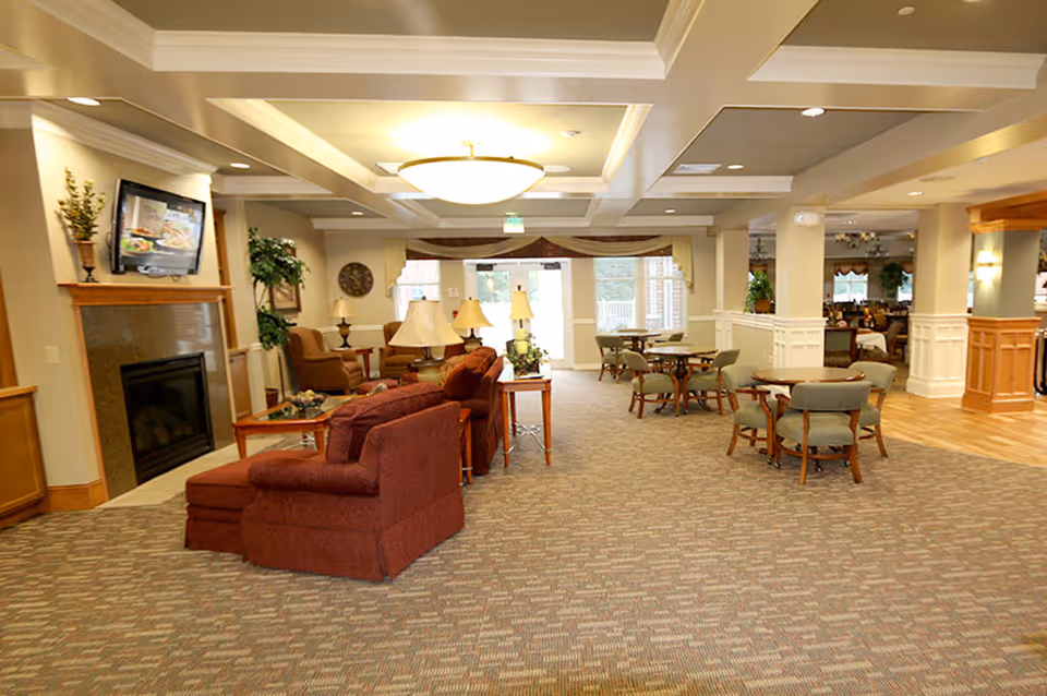 Spacious senior living common area with sofas, armchairs, small dining tables, a fireplace and a wall-mounted TV.