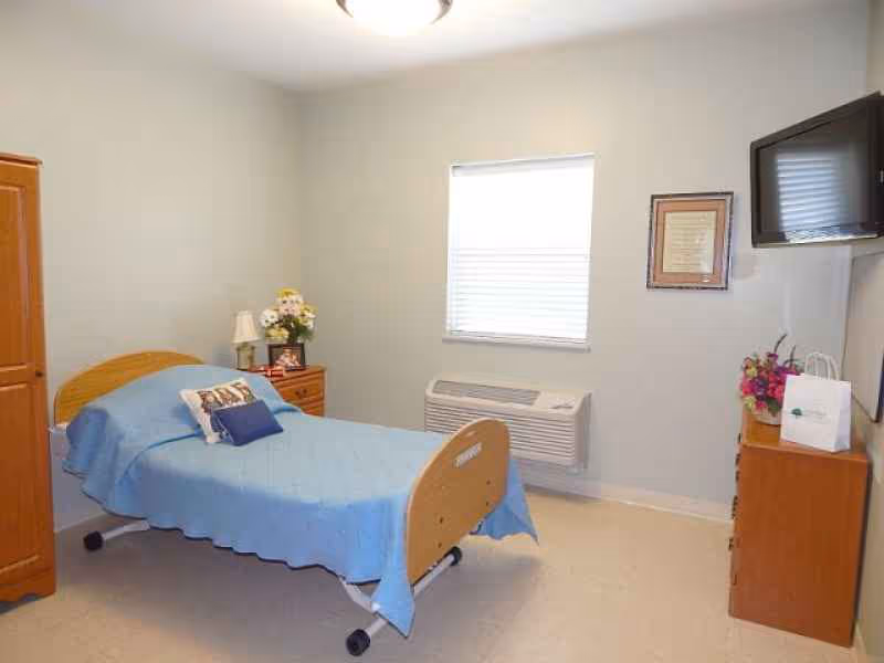 A simple, clean bedroom with a single bed covered in a blue blanket and two pillows. There is a wooden nightstand with a lamp and a vase of flowers next to the bed. A wooden dresser with a bouquet of flowers and a small white bag is against the wall. A flat-screen TV is mounted on the wall above the dresser. A window with blinds is centered on the far wall above a wall-mounted air conditioning unit. The walls are painted light gray.