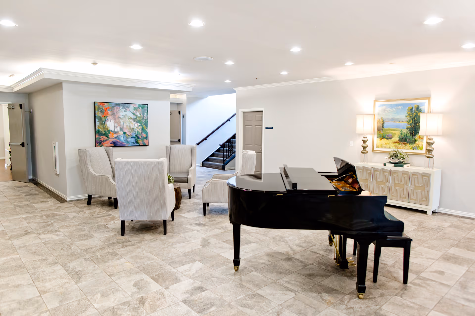 Bright lounge with a black grand piano, a cluster of upholstered chairs, artwork, and a sideboard in a senior living common area.